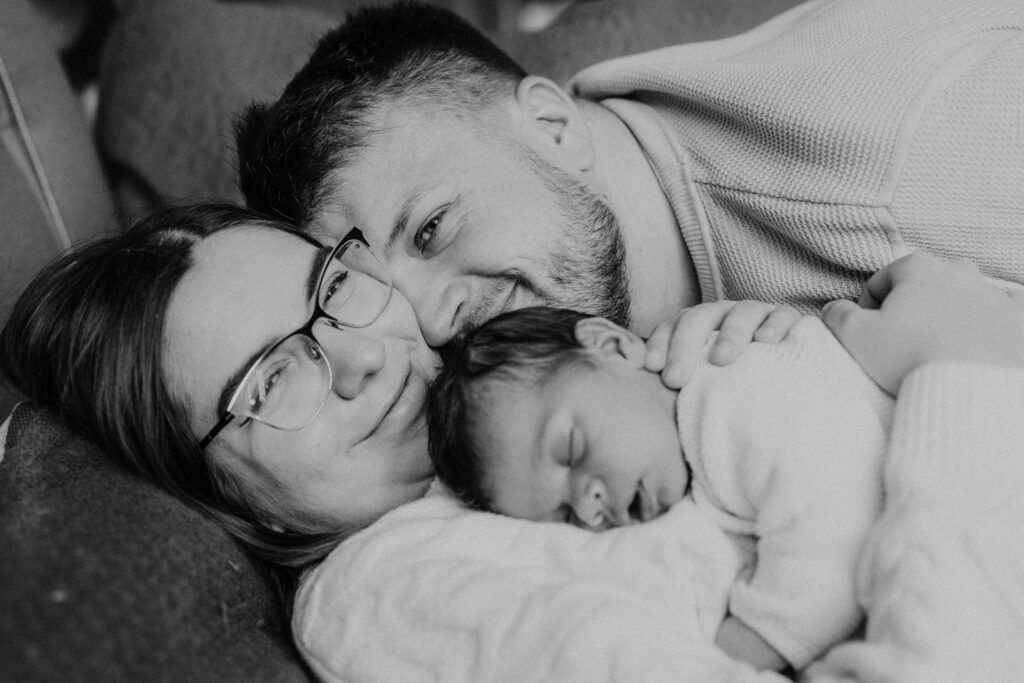 Uśmiechnięcie rodzice przytulajs sie do siebie leżąc obok swojego 2 tygodniowego nowowrodka.