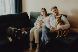 Rodzice ze swoim 2 tygodniowym synkiem siedzą na kanapie w sowim domu a obok nich siędzą ich 2 psy.