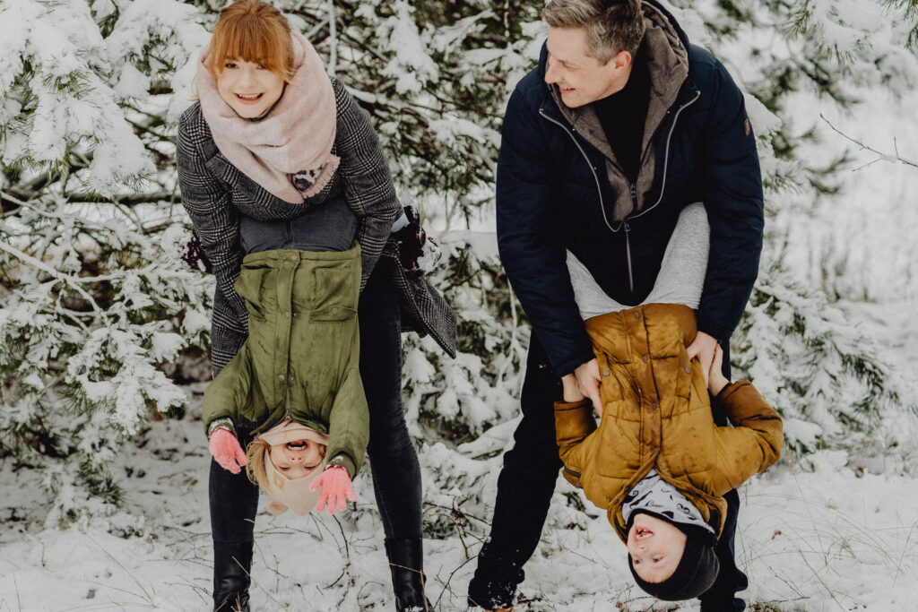 Rodzice trzymają dzieci głowami do dołu podczas fotograficznej sesji lifestyle w Białymstoku. Jest zima, wszędzie śnieg a dzieci są w czapkach.