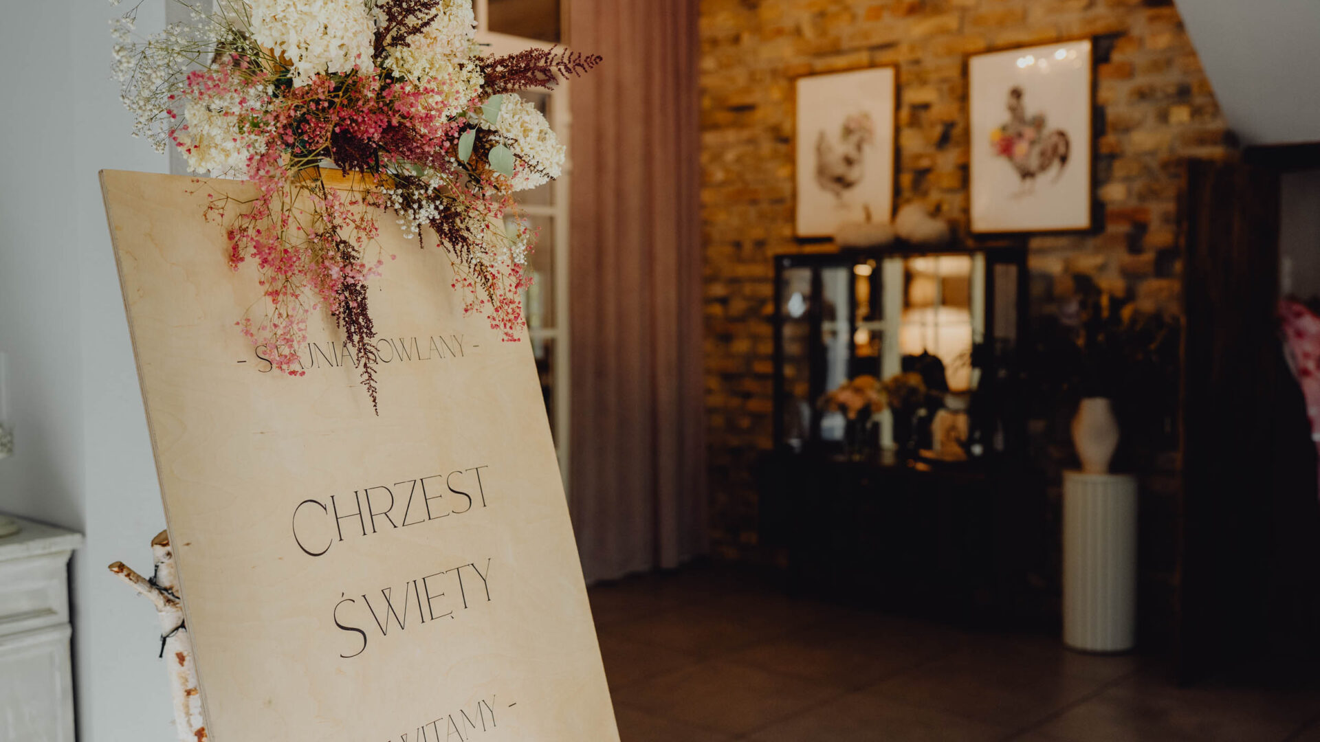 Stojący na wejściu udekorowany kwiatami witacz z napisaem zapraszamy na Chrzest Święty.