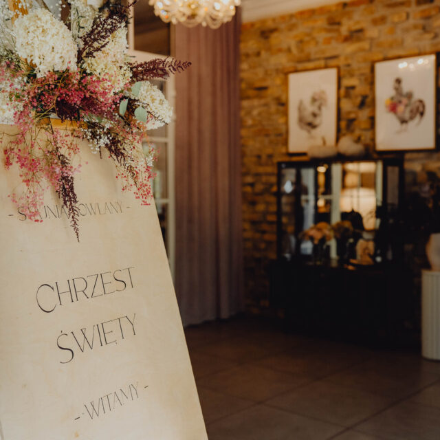 Stojący na wejściu udekorowany kwiatami witacz z napisaem zapraszamy na Chrzest Święty.