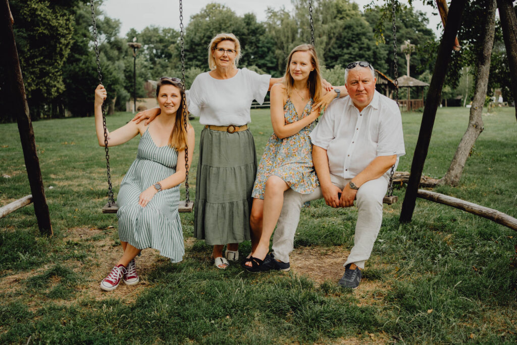 Dziadkowie pozują do zdjęcia na fotograficznej sesji pokoleniowych wraz z swoimi dorosłymi córkami. Dziadkowie siedzą na huśtawce a córki stoją przy nich.