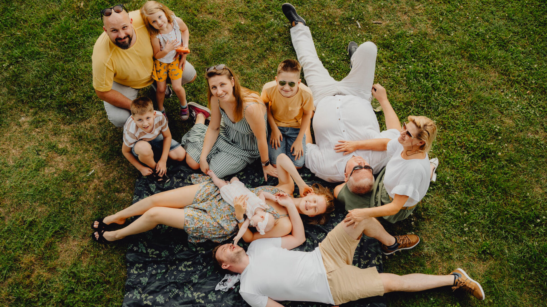 Duża rodzina pozuje do zdjęcia na sesji pokoleniowej siedząc na kocu na łące. 10 osób uśmiecha się i patrzy w obiektyw. Ludzie obejmują się.