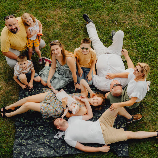 Duża rodzina pozuje do zdjęcia na sesji pokoleniowej siedząc na kocu na łące. 10 osób uśmiecha się i patrzy w obiektyw. Ludzie obejmują się.