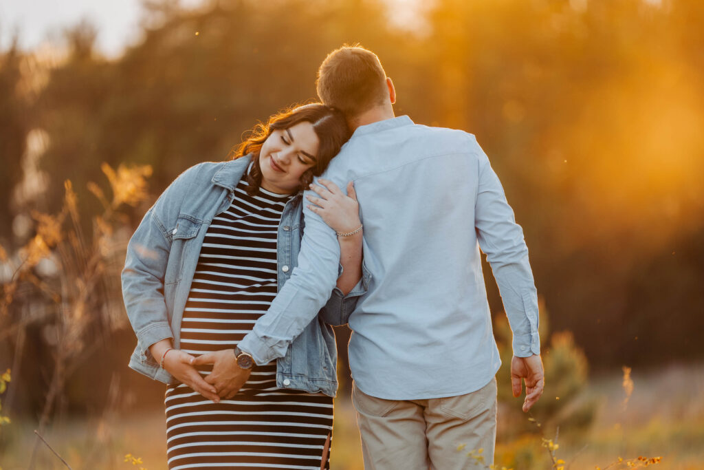 Para przytula się ramie w ramie na sesji ciążowej. Jest złota godzina, zachód słońca w tle. Mężczyzna trzyma dłoń na brzuchu kobiety.
