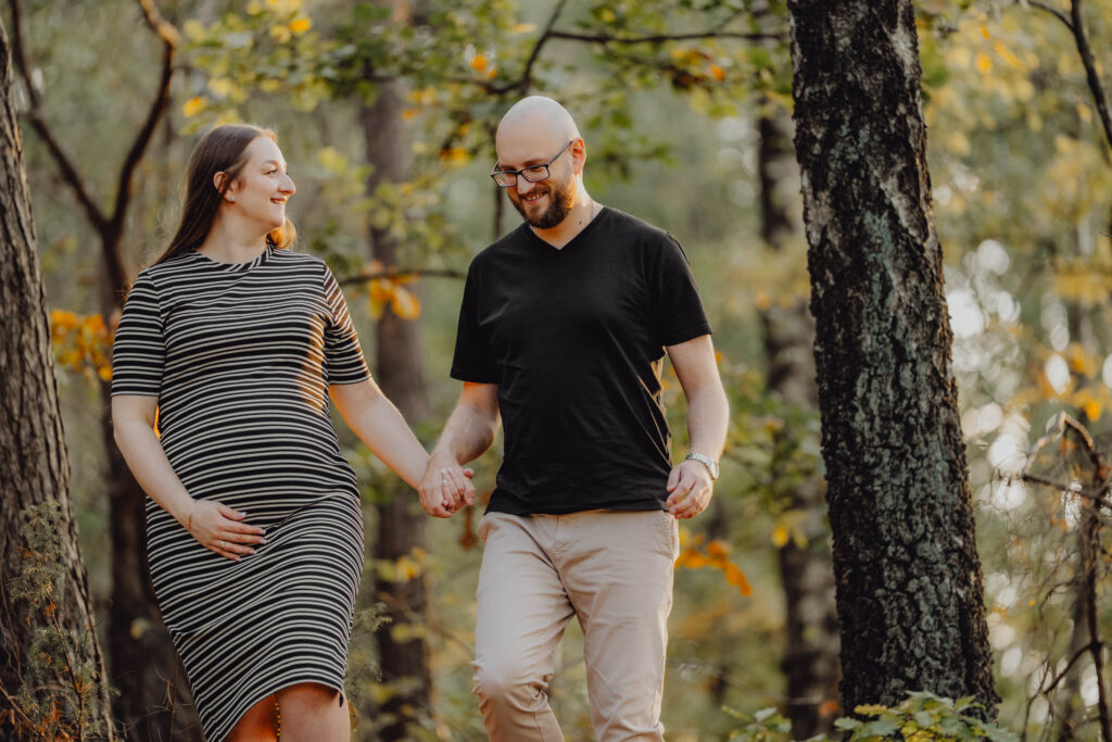 Para na sesji ciążowej w lesie, idzie trzymając się za ręce patrzy sobie w oczy. Są uśmiechnięci.
