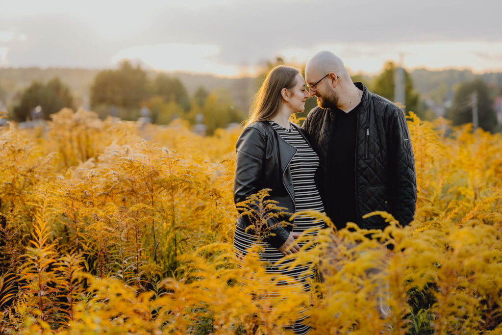 Para przytula się czołami na sesji ciążowej w plenerze. Jest kolorowo, w tle zachód słońca.