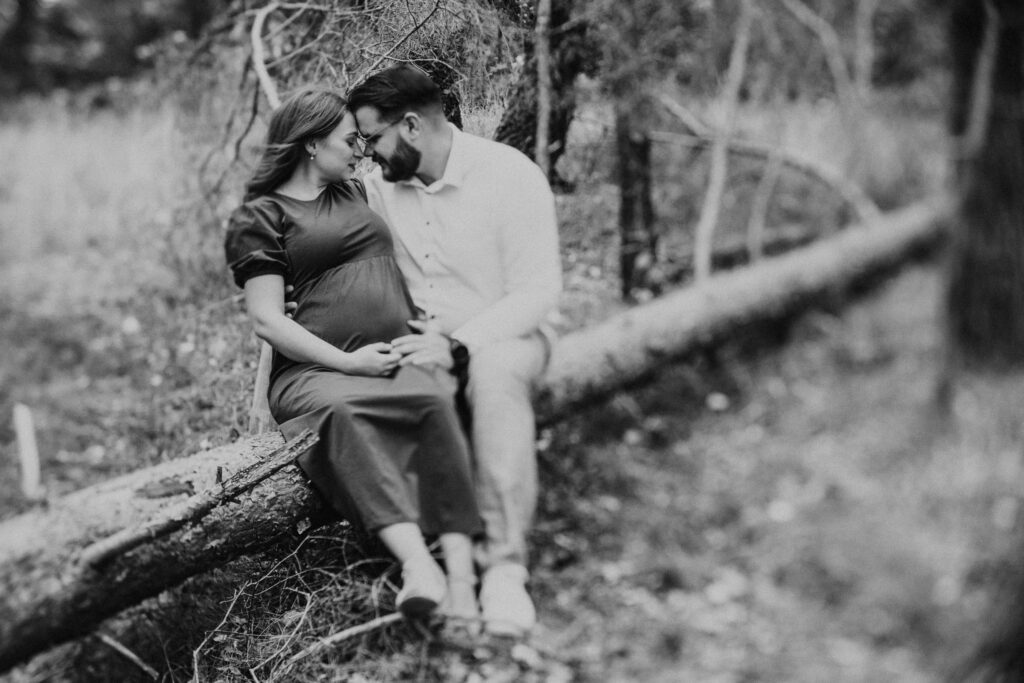 Artystyczne czarno białe zdjęcie przytulnej pary na sesji ciążowej. Para siedzi na zwalonym pniu drzewa. Poza nimi wszystko jest rozmyte.