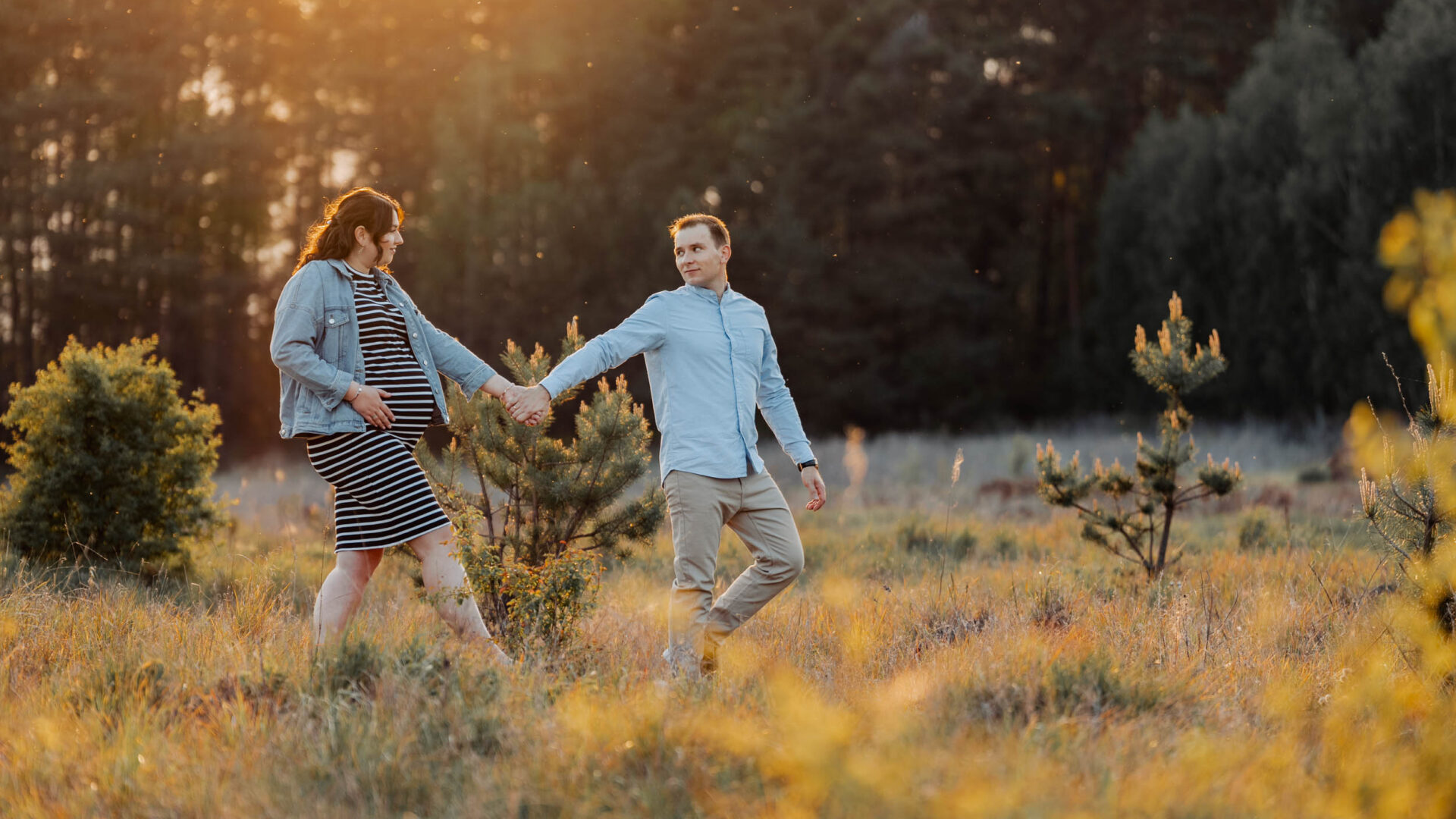 Mężczyzna prowadzi za rękę swoją ciężarną żonę na łące podczas sesji ciążowej. Jest zachód słońca.