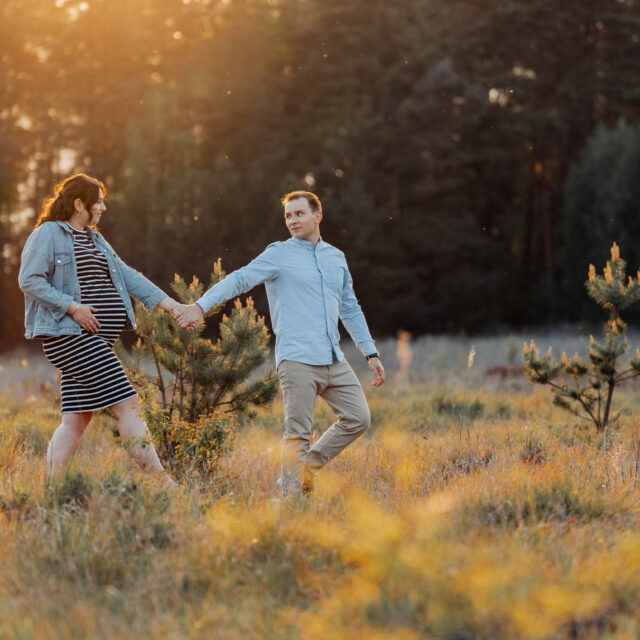 Mężczyzna prowadzi za rękę swoją ciężarną żonę na łące podczas sesji ciążowej. Jest zachód słońca.